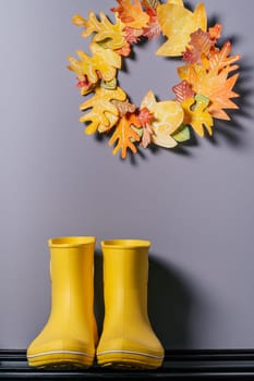 Homemade autumn yellow-red paper wreath and yellow rubber boots on gray wall background. Seasonal DIY. Vertical