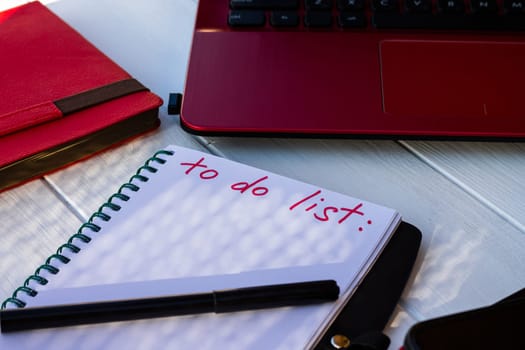 Notebook with To Do List with phone, red diary and laptop on a white wooden background