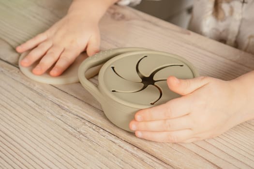 Unrecognizable child holding pastel gray silicone snack cup near lid at wooden table. Baby accessories, tableware, first feeding concept