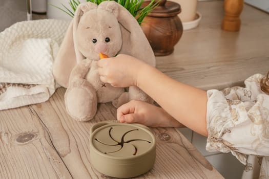 Unrecognizable child taking piece of carrot out from pastel gray silicone snack cup and feeding toy bunny at wooden table. Baby accessories, tableware, first feeding concept