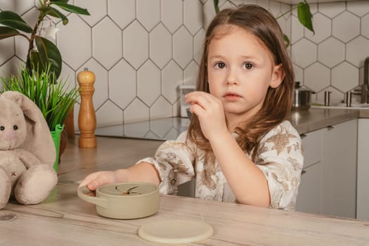 Little dark haired girl 3-4 years old taking piece of food out from pastel gray silicone snack cup and looking into frame in kitchen. Toy bunny nearby. Baby accessories, tableware, feeding concept