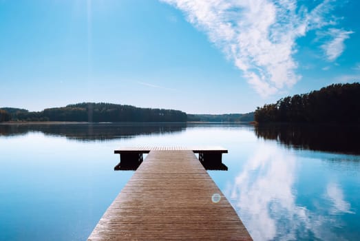 A wooden pier against the backdrop of a lake in the middle of the forest. Blue sky with clouds reflected in the water of Lake Baltieji Lakajai in Labanoras Regional Park.