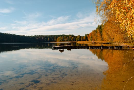 Lake Baltieji Lakajai with clear water in Labanoras Regional Park and a wooden marina with benches for relaxation. Autumn trees are covered in the water