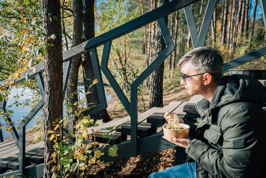A man sits on the shore of Lake Baltieji Lakajai in Labanoras Regional Park, Lithuania in a warm kurta and eats a salad of disposable paper dishes while relaxing