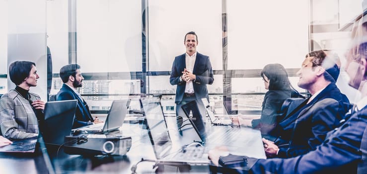 Successful team leader and business owner leading informal in-house business meeting. Business people working on laptops in foreground and glass reflections. Business and entrepreneurship concept.