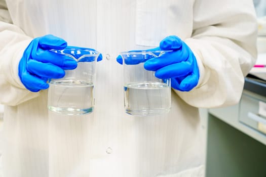 A person in a lab coat is holding two beakers with clear liquids