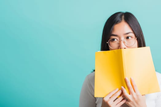 Young beautiful Asian woman hiding behind an open book, Portrait female in glasses is holding and reading a book, studio shot isolated on a blue background, Education concept