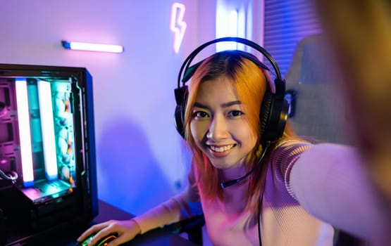 Live stream. Asian woman wearing gaming headphones at home, Happy gamer recording and saying hello to her online fans during live streaming before playing video game online with computer neon lights