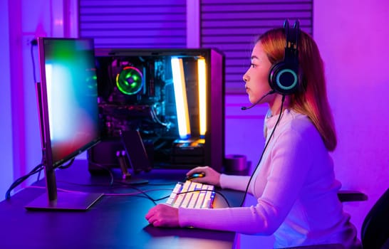 Asian professional gamer playing online video game on desktop computer PC have colorful neon LED lights, young woman in gaming headphones using computer for playing game at home