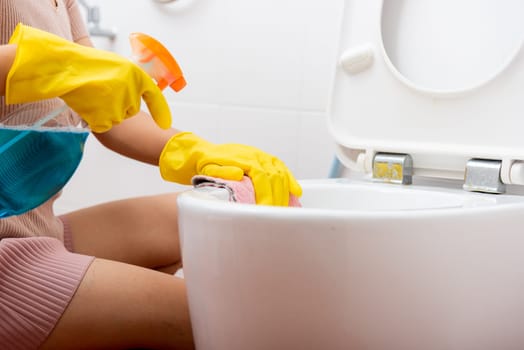 Hand of Asian woman cleaning toilet seat using liquid spray and pink cloth wipe restroom at house, female wearing yellow rubber gloves she sitting cleanup bowl bathroom, Housekeeper healthcare concept