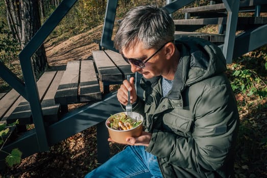 A man dines on a salad from a disposable paper bowl in the open air during a walk in nature in the fall.