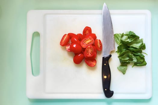 High angle shot of kitchen knife resting on white chopping board with cherry tomatoes in half and fresh basil leaves.