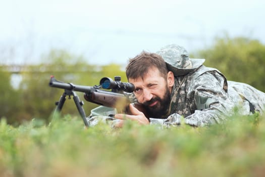 The hunter is aiming and shooting with sniper on shotgun, lying down on grass.