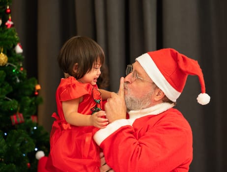 Santa Claus wearing red costume keeping forefinger by his mouth while trying to comfort shouting crying toddler girl near Christmas tree in living room at home
