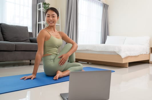 Young attractive woman practicing yoga, doing Half lord of the fishes exercise, Ardha Matsyendrasana pose. Asian woman sitting on blue mat and looking at laptop for practice yoga online course at home. Healthy and sport lifestyle.