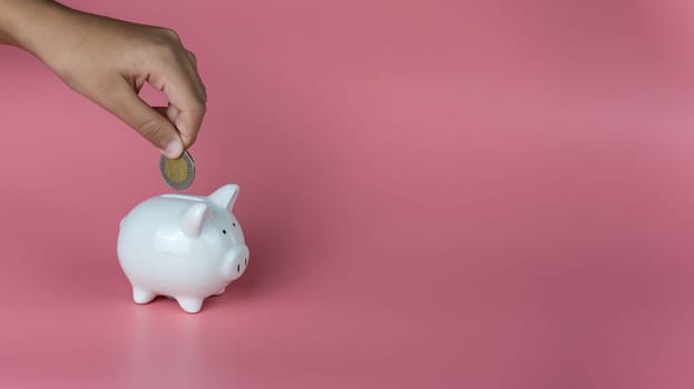 A human hand put a coin in a piggy bank on a pink background. Savings and investment concepts.