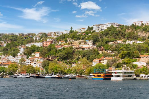 View from the sea of the green mountains of the Europian side of Bosphorus strait, with docked boats, traditional houses and dense trees in a summer day