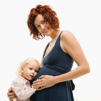 Caucasion mid-adult attractive pregnant smiling woman kneeling in front of female toddler who is pressing ear against belly.