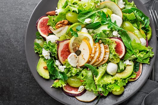 Fresh green leafy salad with mozzarella cheese, fruits and nuts on dark background close up. Concept for a tasty and healthy meal