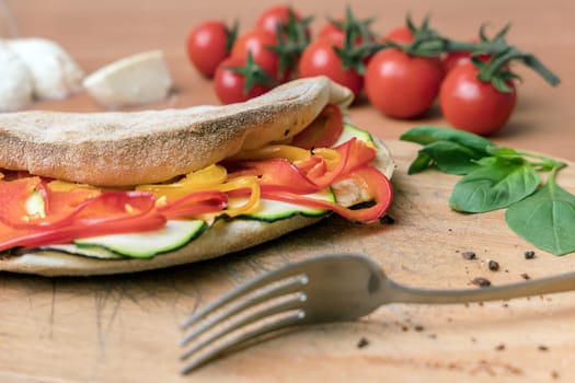 Italian food, closed vegetarian calzone pizza on rustic wooden background. Peppers and zucchini. On background small red tomatoes, mozzarella and basil. Fork. Selective focus.