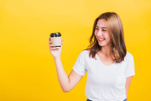 Portrait Asian Thai beautiful happy young woman wear white t-shirt standing smiling holding take away coffee paper cup, studio shot isolated on yellow background, with copy space