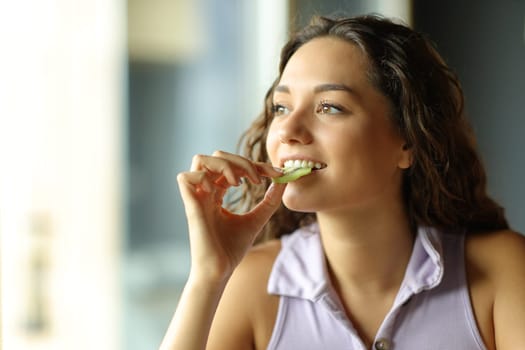 Happy woman eating kiwi at home looking through a window