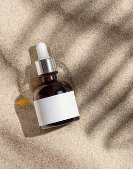 Dropper spray bottle with blank label on beige sand top view. Packaging mockup. Natural skincare product for beauty routine. Summer sunny day with palm leaf hard shadow, tropical vacation mood