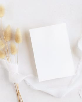 Blank card near silk ribbons and dried hare's tail grass top view on white, mockup. Boho romantic scene with vertical card. Bohemian or close to nature greeting or wedding Invitation
