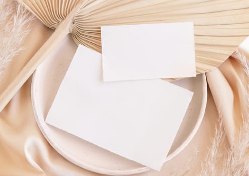 Blank cards on plate with dried palm leaf and beige silky fabric close up, mockup. Romantic scene with horizontal paper card. Bohemian pastel greeting or wedding Invitation .