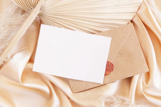 Card and sealed envelope on beige satin fabric and dried palm leaf close up, mockup. Romantic Boho scene with horizontal paper card and envelope. Bohemian or close to nature greeting or wedding Invitation.