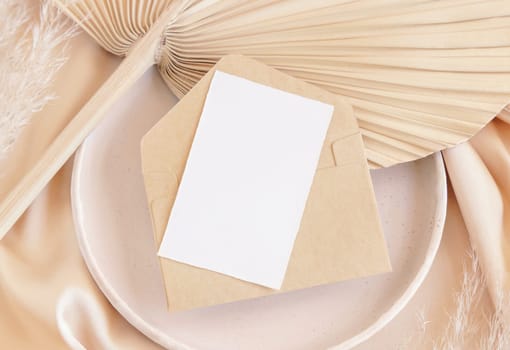 Blank card and envelope on plate with dried palm leaf and beige silky fabric close up, mockup. Romantic scene with vertical paper card. Pastel greeting or wedding stationery.