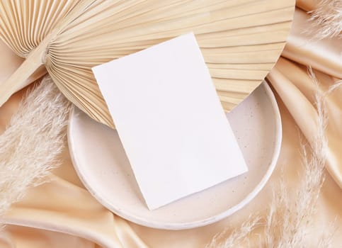 Blank card on plate with dried palm leaf and beige silky fabric close up, mockup. Romantic scene with vertical paper card. Bohemian pastel greeting or wedding Invitation .