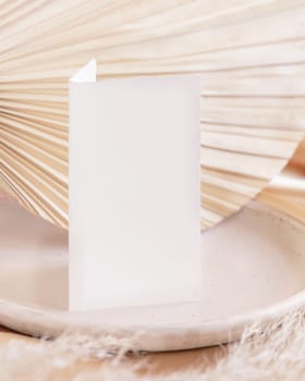 Folded card on plate near dried palm leaf and beige silky fabric close up, table place mockup. Romantic scene with vertical paper card. Pastel greeting or wedding stationery.