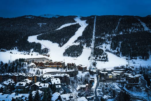 El Tarter village at dusk or night fall with snowy ski slopes cutting through forest hills in Pyrenees mountains, Grandvalira, Andorra