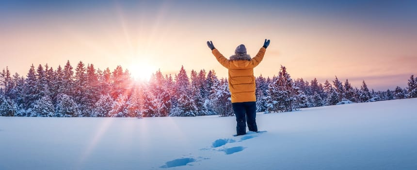 A woman stands on a snowy field with her hands raised up. Panoramic background.