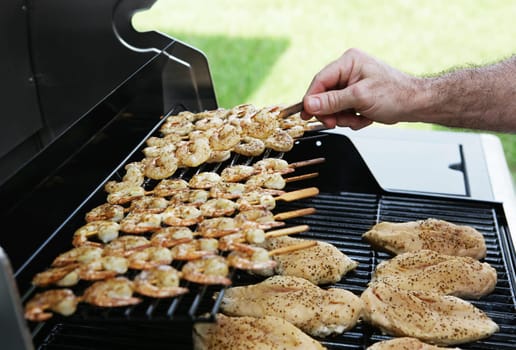 Shrimp and chicken on a barbecue grill.  A man's hand is flipping a skewer of shrimp.  Focus on shrimp being turned.