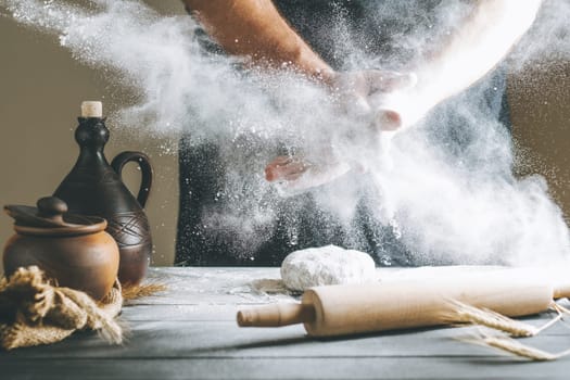 Male hands with flour clap over dough next to clay pot and oil bottle and rolling pin. Flour splash over dark table while cooking