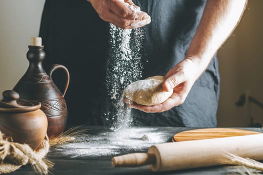 Male hands pour flour on the dough next to clay pot and oil bottle and rolling pin on dark table while cooking