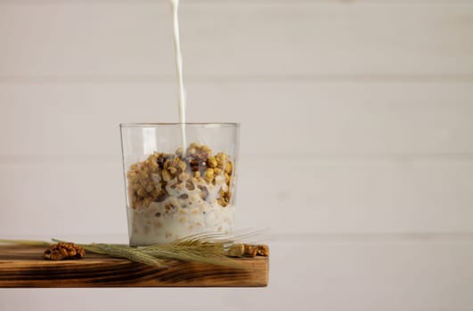 Milk flowing into a cereal granola in a transparent glass with rye spike and walnuts on a shelf on a white background. Copy space.