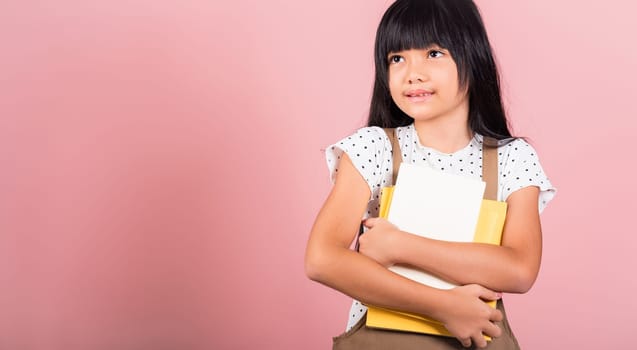 Asian little kid 10 years old holding book for going to school at studio shot isolated on pink background, Happy child girl hugging textbooks dictionary, lifestyle lovely educated cheerful