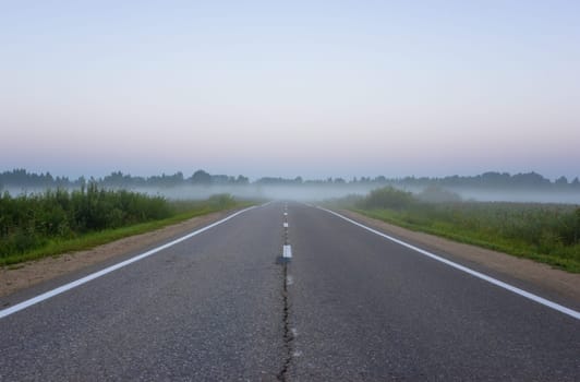 Diminishing perspective view of an empty, straight suburban asphalt roadway with white road markings vanishing into the morning mist at dawn. Yaroslavl Oblast, Russia.