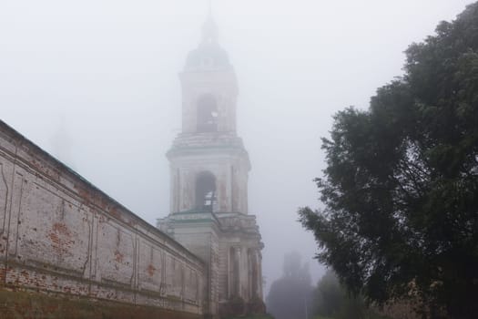 Bell tower and the brick wall of the ancient Spaso-Yakovlevsky Orthodox monastery covered with autumn morning fog. Russian classical architecture of the XVIII century. Rostov Veliky, Yaroslavl oblast.