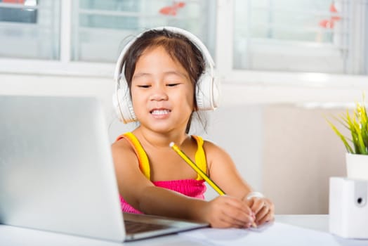 Little kid preschool wear headphone sit at desk use laptop computer and communicates on Internet online video call with teacher, Asian child girl study video conference distant education at home