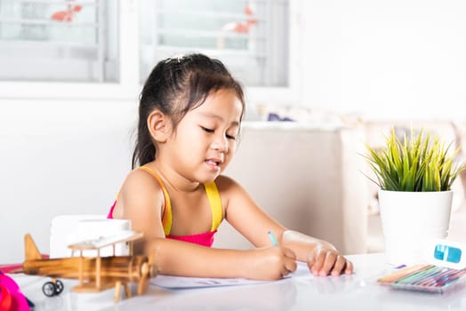Child little girl drawing cartoon on paper before paint the color, Asian cute kid preschooler sitting on table smiling she draw and writing a picture with pencil at home to learning arts homework