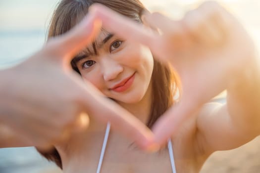 Close up Asian happy young woman looking through making shape camera frame made from her fingers on the beach at sunset, Composition finger frame