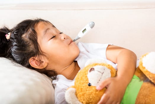 Sick kid. Mother checking temperature of her sick daughter with thermometer in mouth, child laying in bed taking measuring her temperature for fever and illness