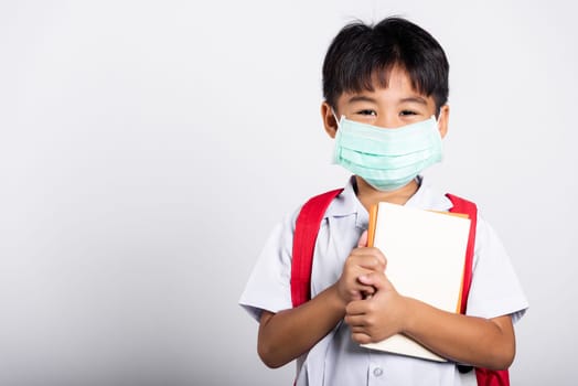 Asian student kid boy wearing student thai uniform and medical protect face mask and hugging note book in studio shot isolated on white background, new normal back to school
