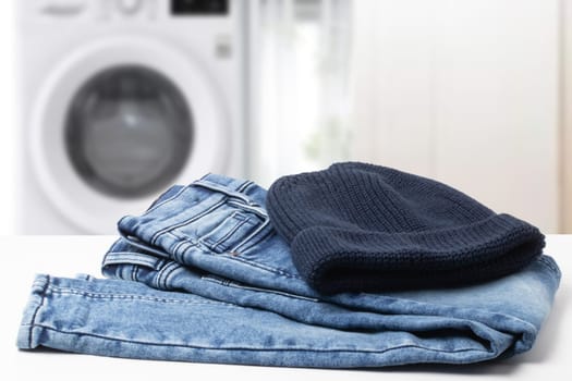 Colorful children jeans clothes. Fashionable folded jeans and a knitted blue Rapper, Beanie or Baseball hat or cap on table over abstract blurred bathroom background.