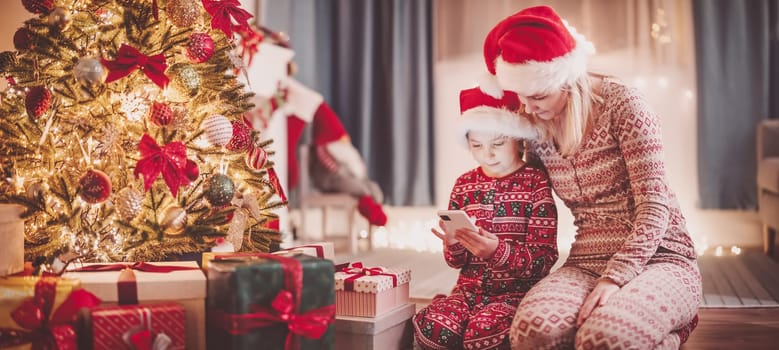 Mother and daughter sitting with smartphone on the floor indoors in Christmas eve. Concept of Christmastime and New Year.