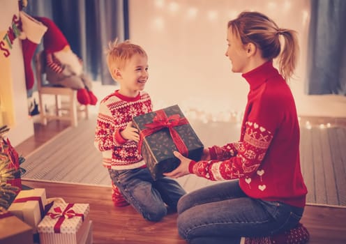 Mother and son indoors sitting on the floor and giving present box each other in Christmas eve. Concept of Christmastime and New Year.